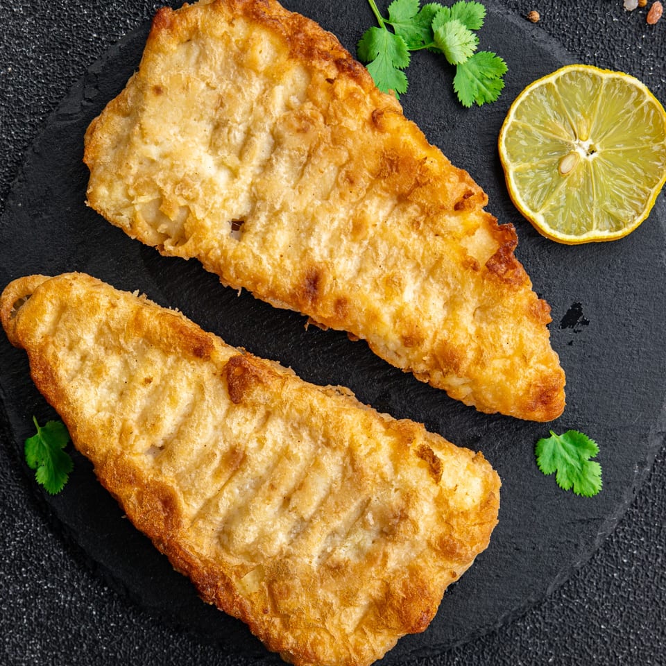 Two golden-brown fried fish fillets with a lemon slice and cilantro on a dark slate.